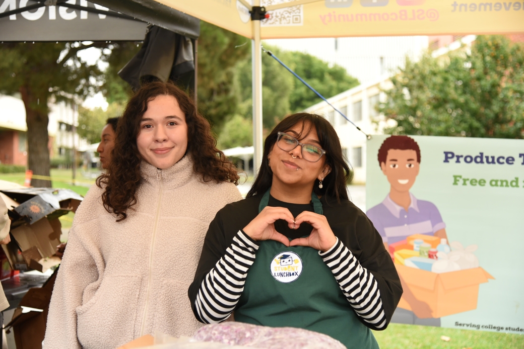 two students standing beside each other, smiling, one of the students is making a heart with her hands.