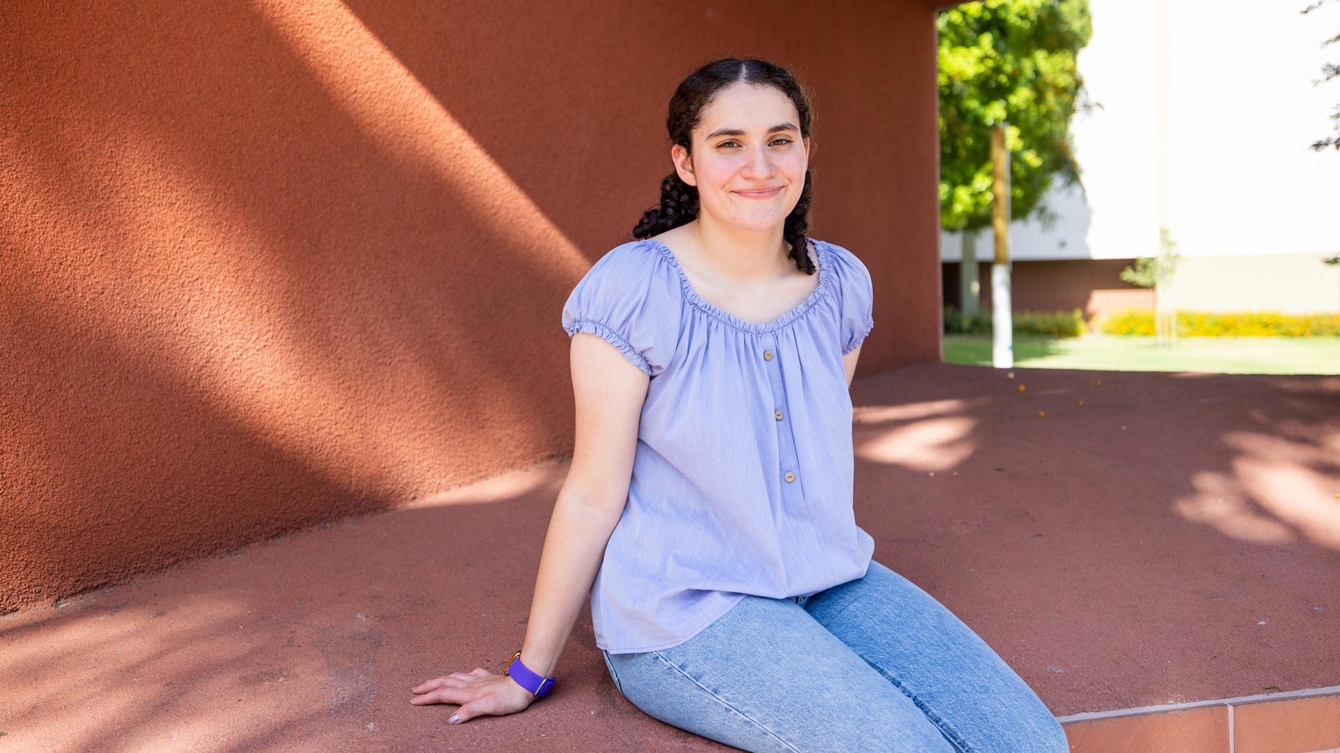 A Cal State LA student smiles while sitting on a bench under a red architectural structure on campus, with trees and lawn in the background.