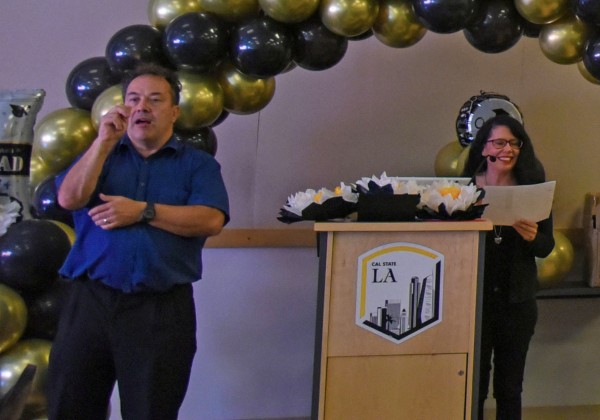 Sign language interpreter standing in the foreground, a person speaking at a podium in the background.