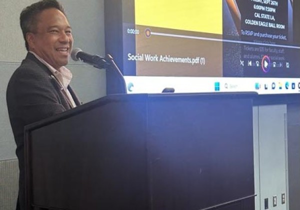 Victor Manalo standing in front of a podium. He is smiling and there is a microphone near his mouth. In the background, there is a projector screen with the image of the inaugural invitation flyer.