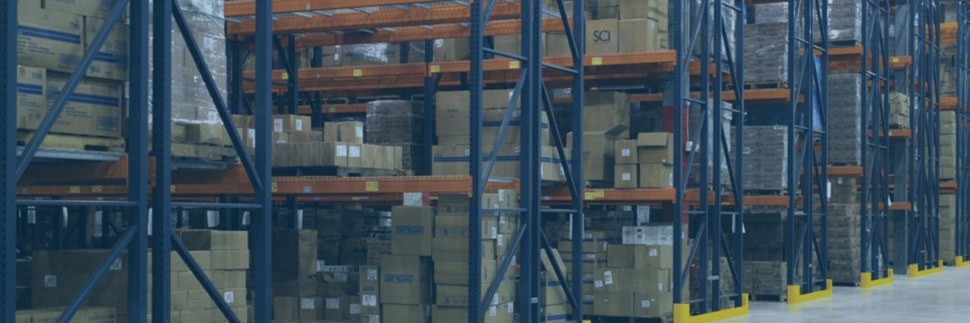 Interior of a warehouse with tall shelving units filled with stacked boxes and pallets along wide aisles.