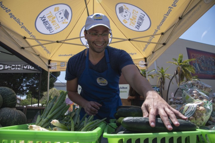 A person standing behind bins of vegetables arranging a cucumber. They are smiling.