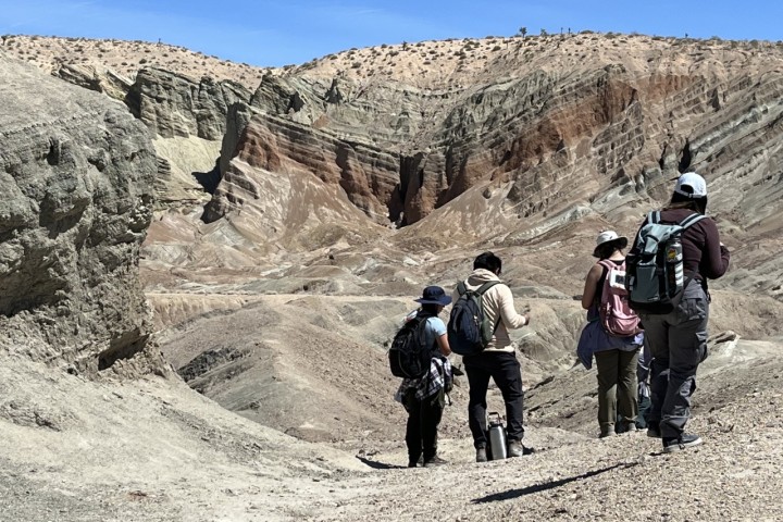 Five students wearing backpacks and hats walk through a rocky desert landscape with layered sedimentary cliffs in the background. The terrain is arid, with light-colored soil and sparse vegetation under a clear blue sky