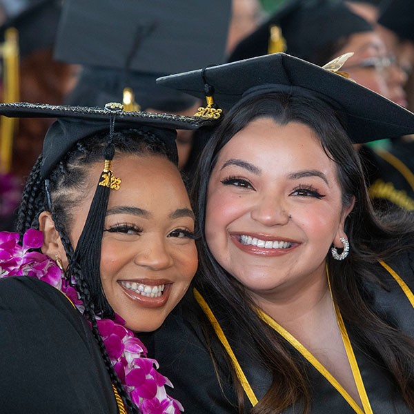 2 graduates at commencement, sitting cheek-to-cheek