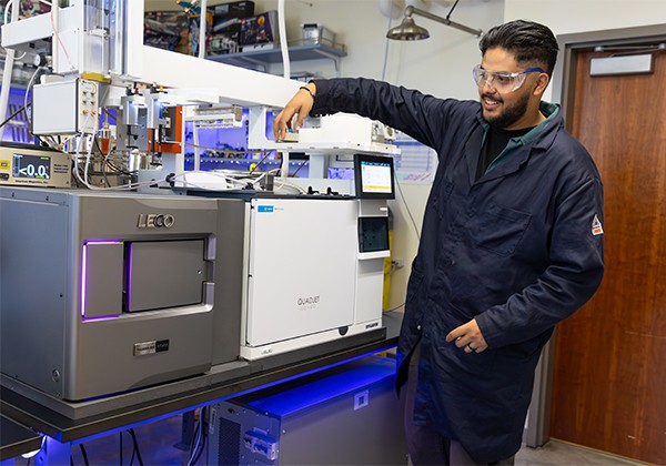 A Cal State LA student wearing a lab coat and safety glasses operates the LECO Pegasus BTX mass spectrometry system inside the LECO Complex Chemical Compositional Analysis Lab.