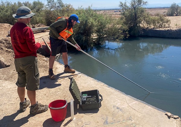 Two researchers collect water samples from a canal using a long pole. They stand on a concrete embankment beside monitoring equipment and a red bucket under bright desert sunlight.