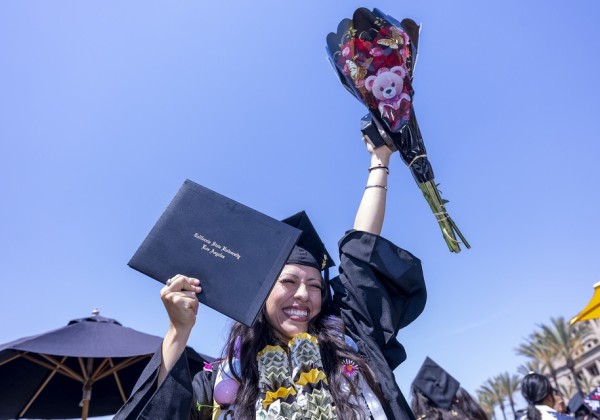 Graduate celebrates outdoors by holding up a diploma cover and bouquet while wearing graduation regalia and celebratory leis at Cal State LA.