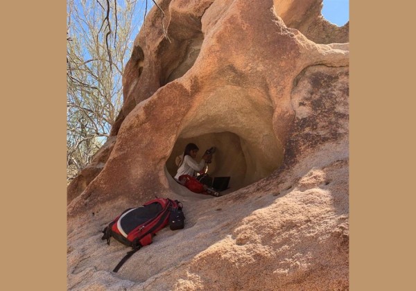 A field researcher works inside a small cave-like rock formation, with equipment and a backpack, surrounded by sunlit boulders and desert vegetation.