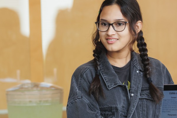 A student with braided hair and glasses holds up a tablet displaying information about garden plants, with refreshments in the background.