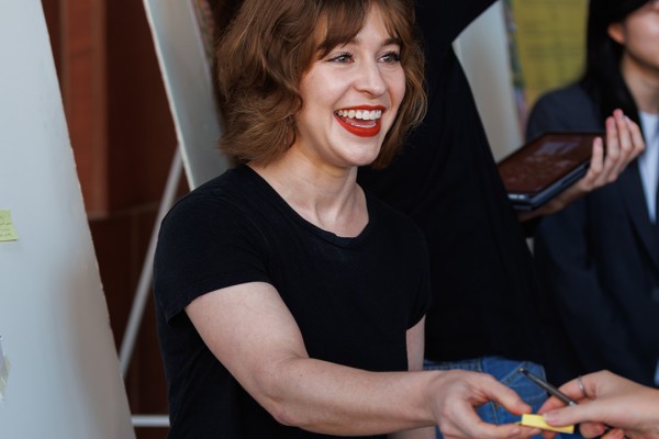 A woman with short hair and a big smile hands out material to an attendee, with a poster in the background.