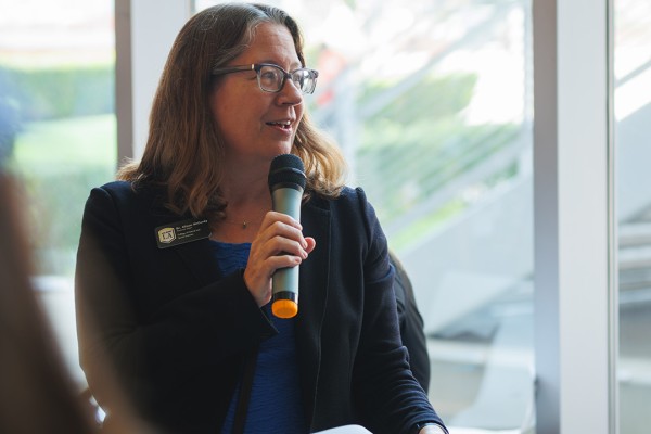 A woman with glasses, wearing a name tag, speaks into a microphone, likely addressing the audience.