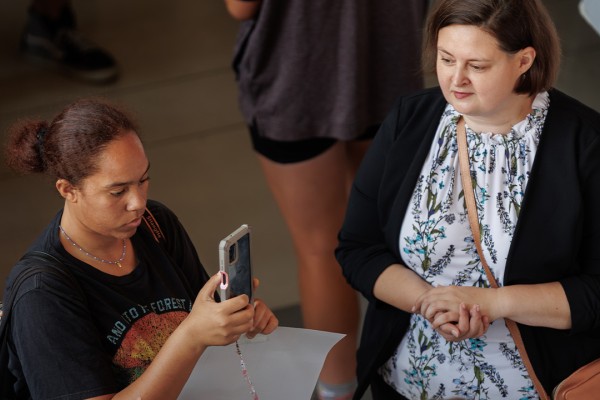 Two women observe event materials, with one taking a photo and the other looking on with interest.