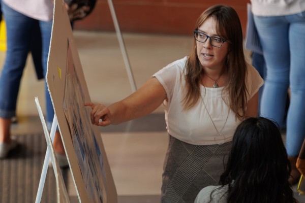 A woman points to information on a large poster board while talking to a small group of attendees.