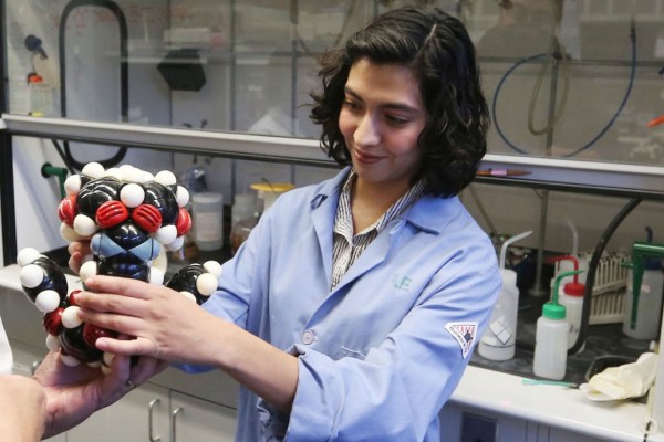 Image of a student in a blue lab coat presenting a model of an atom to a group of fellow students. 