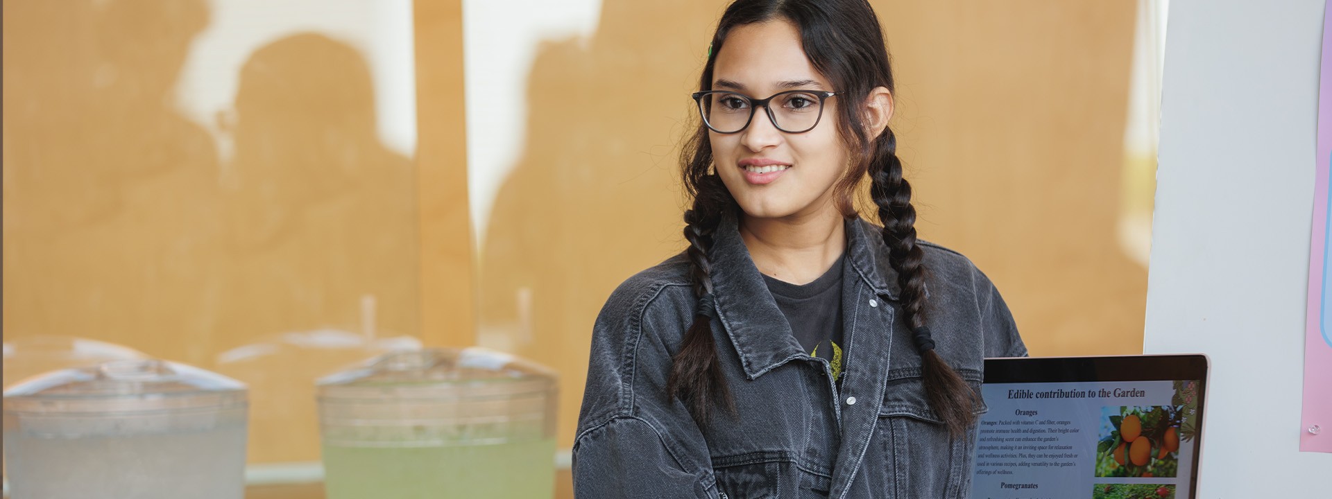 A student with braided hair and glasses holds up a tablet displaying information about garden plants, with refreshments in the background.