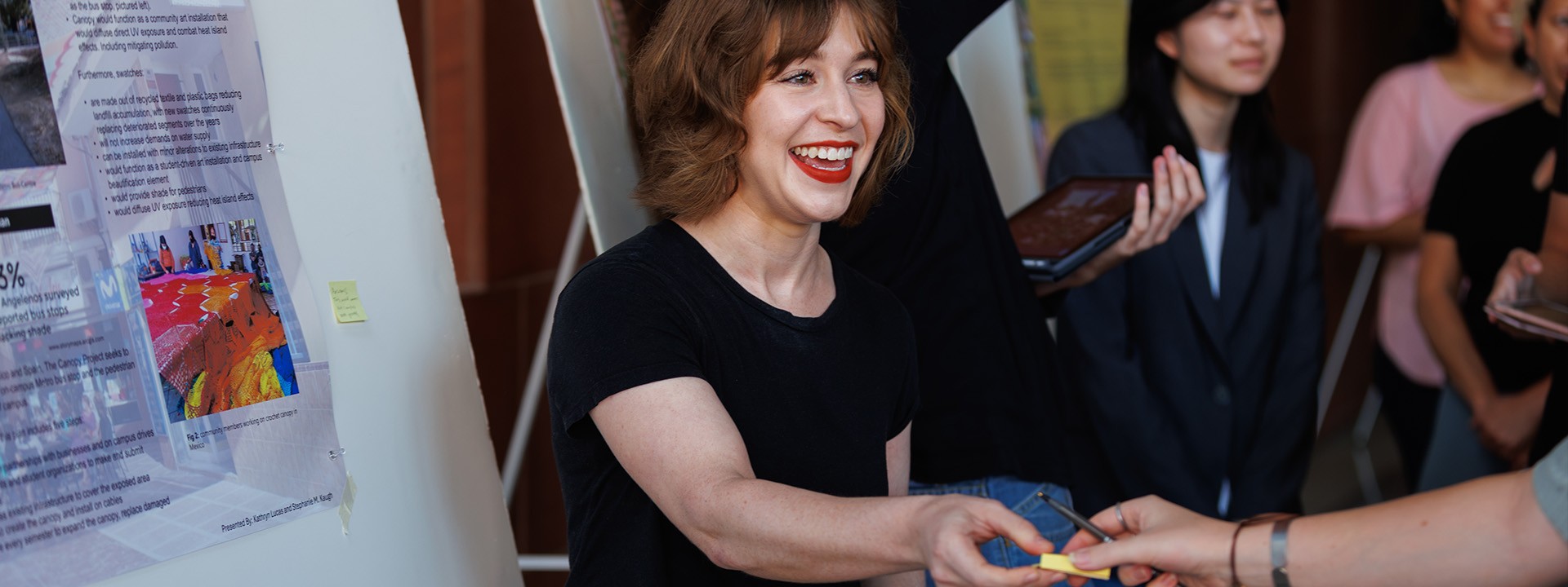 A woman with short hair and a big smile hands out material to an attendee, with a poster in the background.