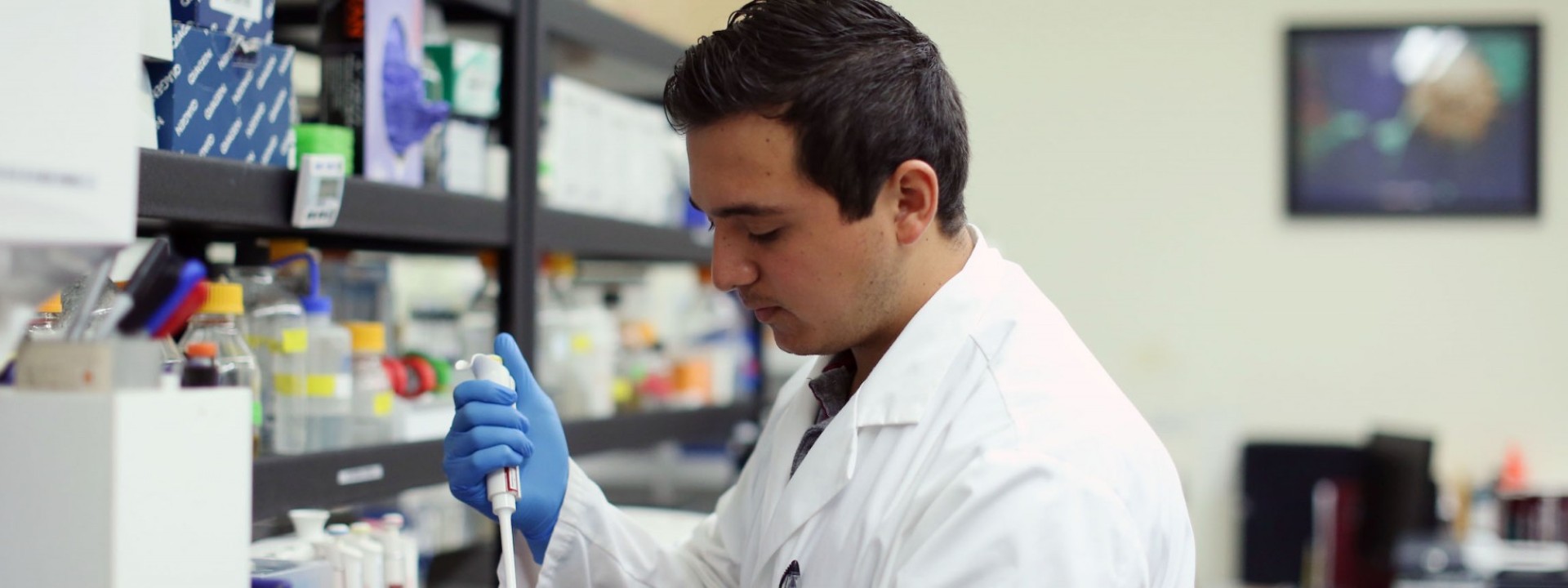 Image of the side profile of a student in a while lab coat and blue gloves using a lab instrument. 