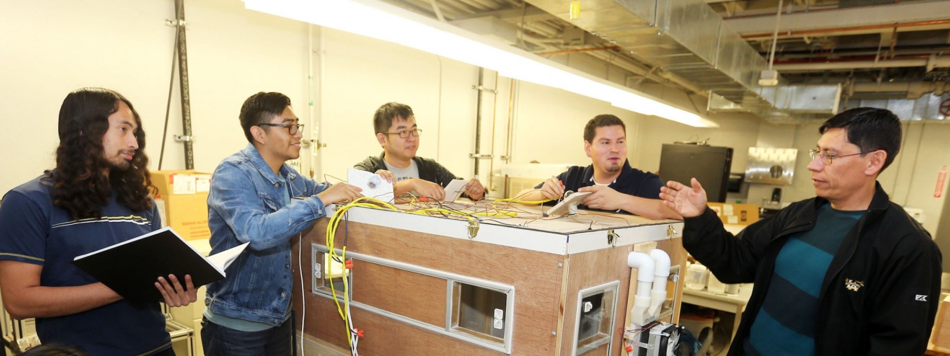 Image of a group of engineering students surrounding a large wooden box device while their professor addresses them. 