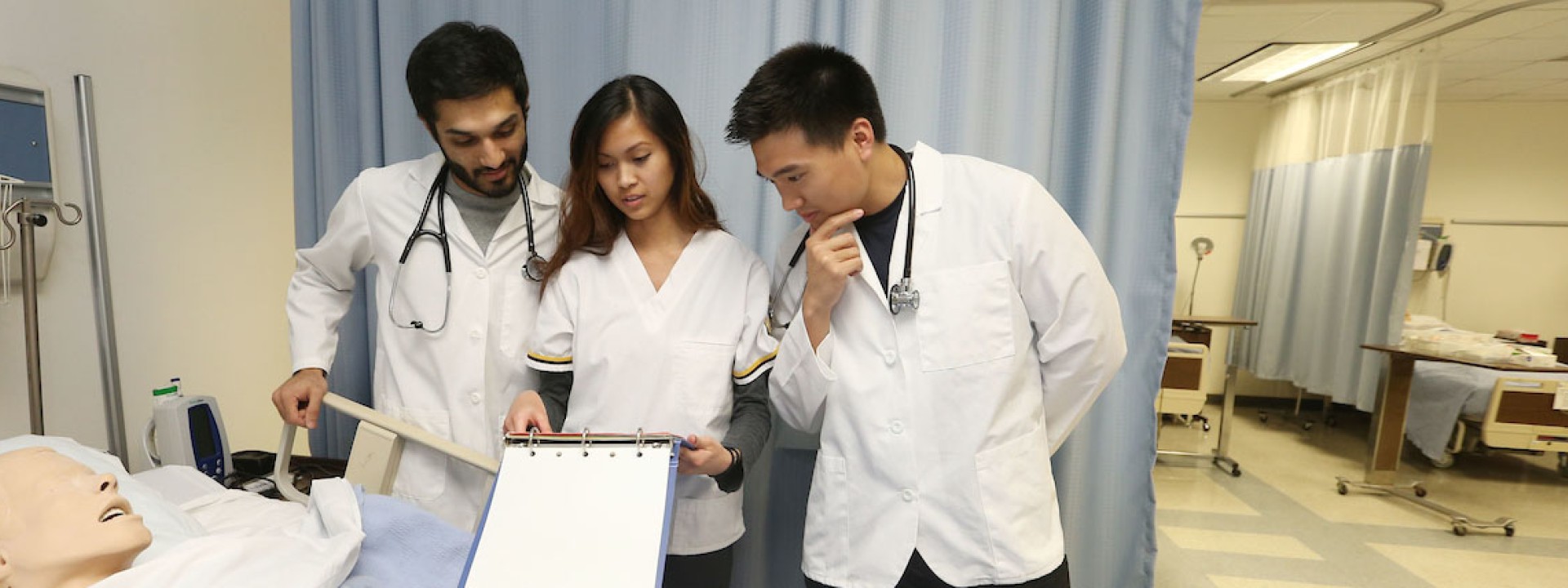 Image of three students in lab coats standing over an educational dummy. 