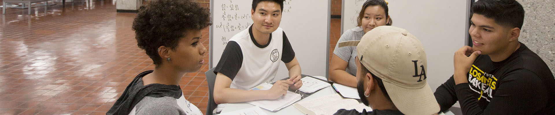 Five students sit around a table in a campus study area, engaged in a group discussion. One student speaks while the others listen and look toward her. Papers, notebooks, and a whiteboard with handwritten notes are visible in the background.